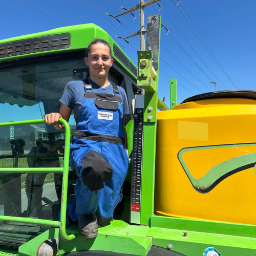 Frau in blauer Arbeitskleidung steht auf einem grünen Traktor bei sonnigem Wetter auf einem Feldweg.